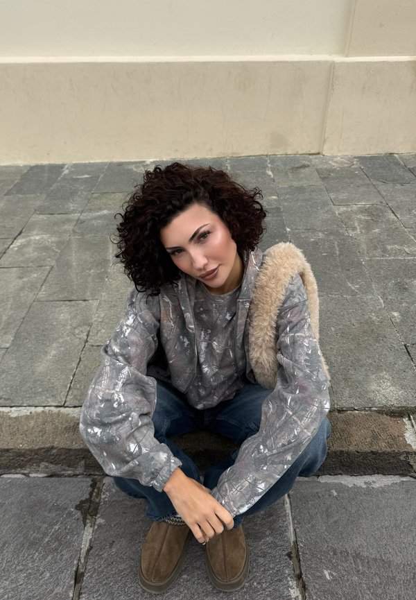 Persona con capelli ricci seduta su un marciapiede di pietra, indossa una felpa con cappuccio grigia a motivi, jeans blu, scarpe marroni e porta una borsa pelosa beige.