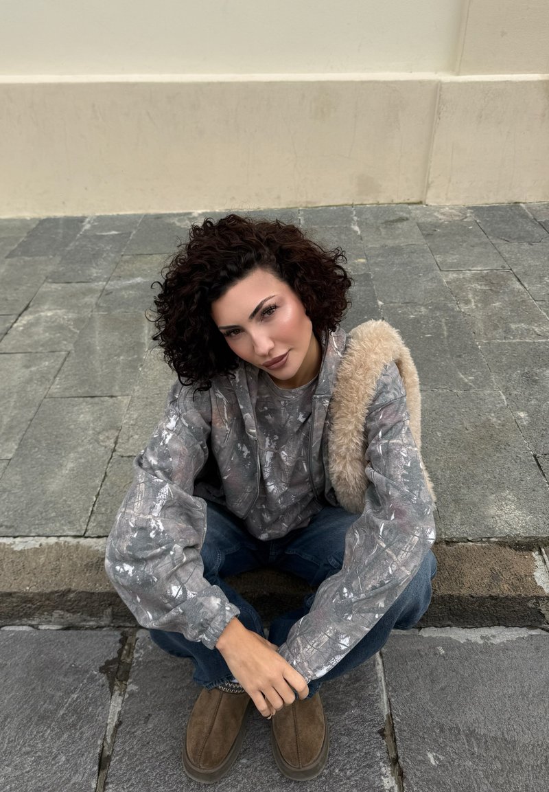 Kiharatukkainen henkilö istuu kivipäällysteisellä kadulla, ja hänellä on päällään harmaa kuvioitu huppari, siniset farkut, ruskeat kengät ja hän kantaa beigeä karvaista laukkua.