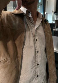 Veste en daim beige avec fermeture éclair et col rabattu, portée sur une chemise à boutons gris clair avec des boutons foncés.
