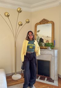 Yellow knitted cardigan, green shirt with "BRASIL" and number 92, black wide trousers, standing beside a decorative lamp and potted plants.