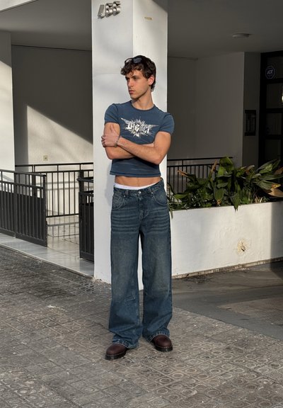 Modelo vistiendo una camiseta azul corta con un diseño gráfico, jeans azules holgados y zapatos marrones, de pie cerca de una pared blanca con plantas.