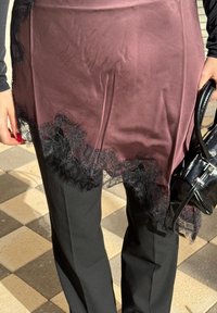 Ruky držiace čiernu kabelku, oblečené v purpurovej saténovej sukni s čiernou čipkou cez čierne nohavice, na podlahe s kockovanými dlaždicami.