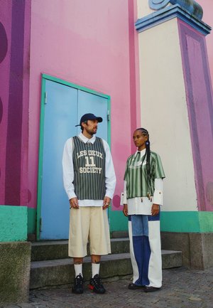 Dos personas están de pie frente a una pared colorida. Uno lleva un chaleco verde a rayas sobre una camiseta blanca y pantalones cortos beige; el otro lleva una camiseta verde a rayas y pantalones vaqueros azules.