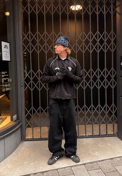 Camisa negra de manga larga con detalles blancos, pantalones negros, guantes y un gorro azul con patrón. De pie frente a una reja de seguridad metálica.