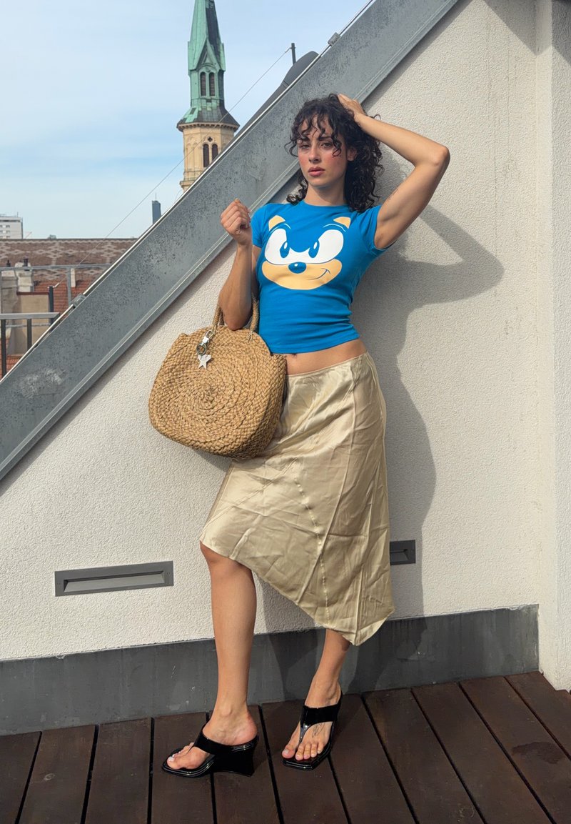 Frau mit lockigem Haar, die ein blaues Sonic the Hedgehog T-Shirt, einen beigen seidigen Rock, schwarze Sandalen mit Absatz trägt und eine große runde gewebte Tasche hält.