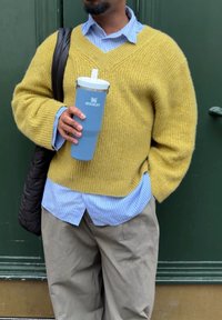 Sennepsgul strikket sweater med V-hals, lagdelt over en lyseblå stribet skjorte. Holder en blå Stanley-termokop. Løse beige bukser.
