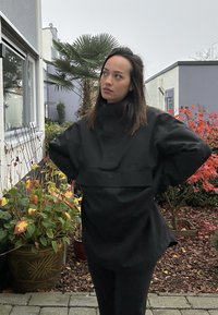 Juoda didelė tunika su aukštu apykakle, plačiomis rankovėmis ir priekyje esantį juostą; lygi tekstūra. Lauko aplinka su augalais ir spalvingomis lapų �šakomis.