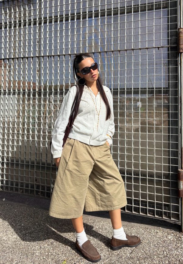 Une femme portant des lunettes de soleil, un chemisier blanc, un short beige ample, des chaussettes et des mocassins pose devant une clôture métallique à mailles, en plein soleil.
