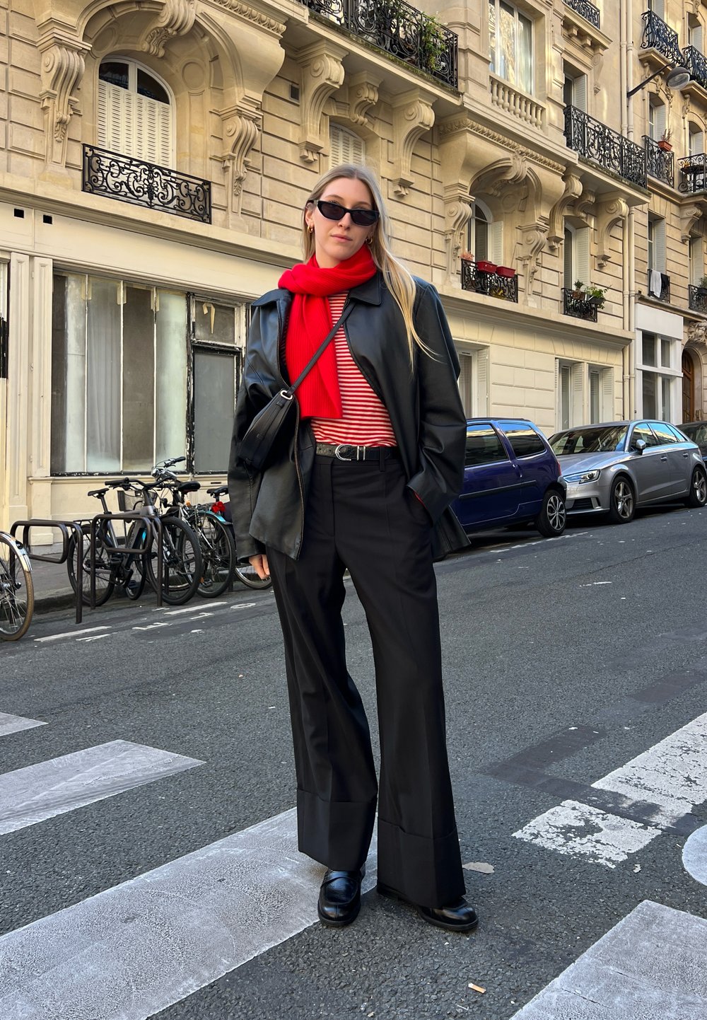 Schwarze Lederoversize-Jacke, roter Schal, gestreiftes Hemd, hoch taillierte schwarze Hose und schwarze Schuhe. Urbane Straßenumgebung mit parkenden Fahrrädern.