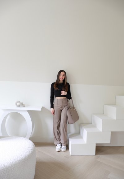 Una persona está de pie junto a una mesa circular blanca y una escalera. Lleva una camiseta de manga larga negra, pantalones de pierna ancha beige y sostiene un bolso topo.