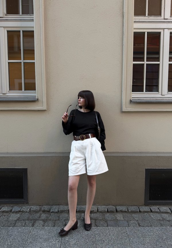 Frau mit dunklem Bob-Haarschnitt, die ein schwarzes Oberteil, weiße Shorts und einen braunen Gürtel trägt, hält eine Sonnenbrille und steht vor einer beige Wand mit zwei Fenstern.