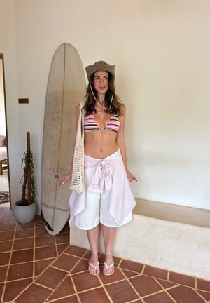 Mujer con top de bikini a rayas, shorts blancos y sombrero ancho, de pie en el interior junto a una tabla de surf y una planta en maceta sobre un suelo de baldosas.
