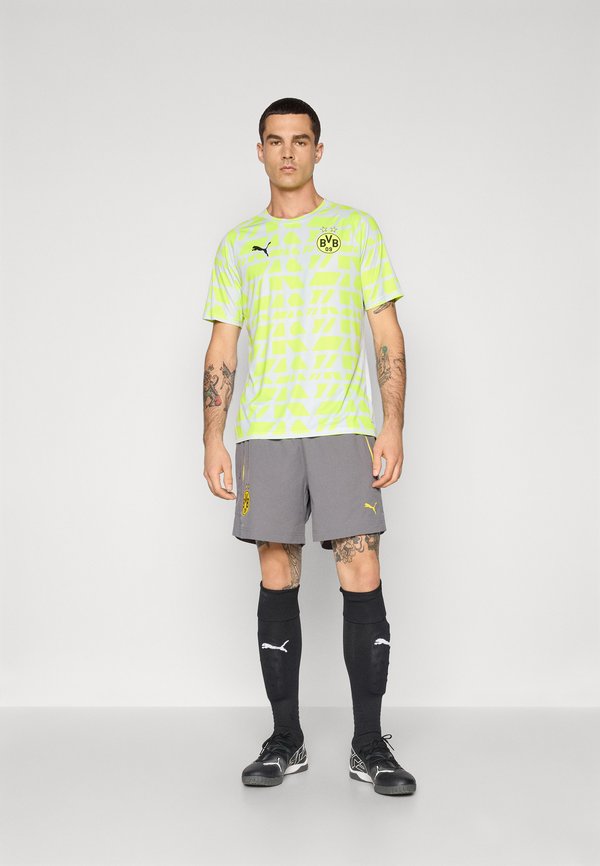 Koszulka sportowa Puma w neonowym żółtym kolorze z geometrycznymi wzorami, połączona z szarymi szortami z żółtym logo, czarnymi skarpetkami i korkami.