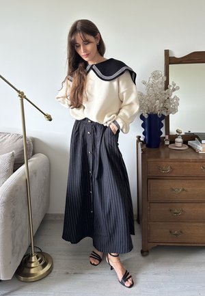 Suéter crema con un cuello marinero negro, combinado con una falda a rayas en blanco y negro con botones y un lazo en la cintura. Sandalias negras con tiras.