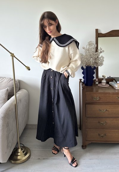 Suéter crema con un cuello marinero negro, combinado con una falda a rayas en blanco y negro con botones y un lazo en la cintura. Sandalias negras con tiras.