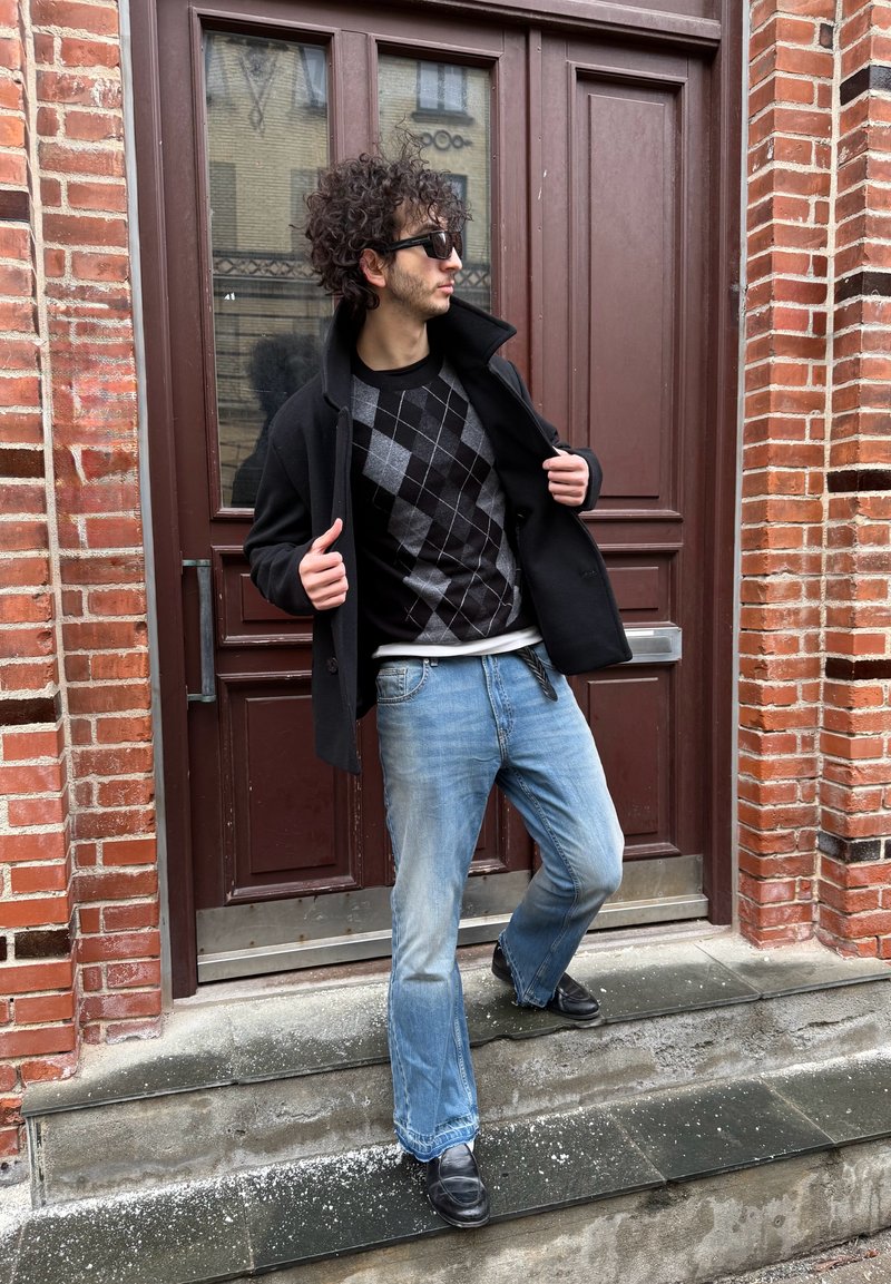 Jeune homme aux cheveux bouclés portant des lunettes de soleil, un manteau noir, un pull à losanges et un jean, debout sur des marches devant une porte en bois marron et un mur en briques.