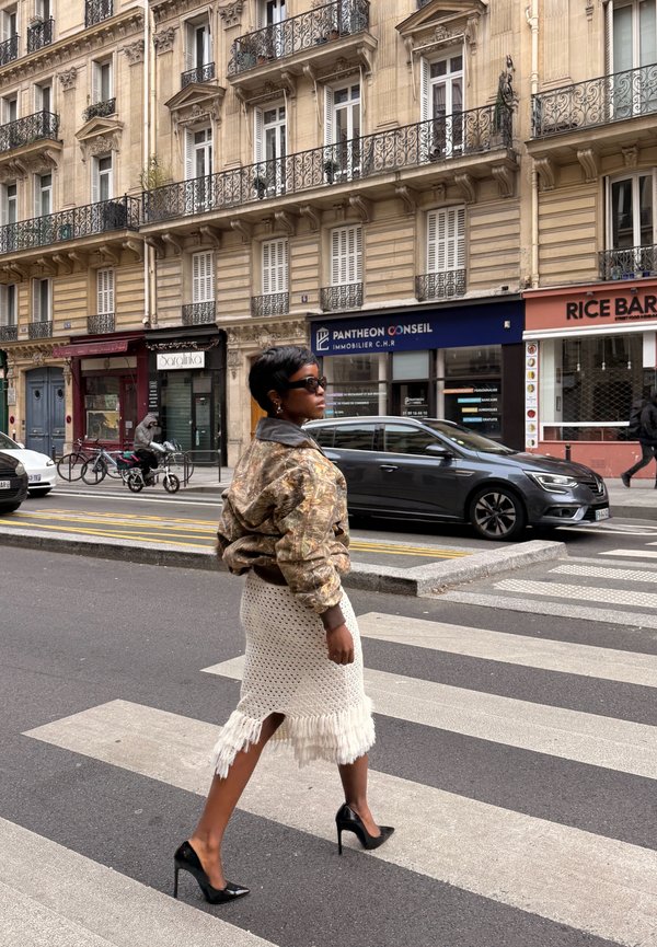 Mujer con gafas de sol, una chaqueta estampada y una falda blanca con flecos caminando por un paso de peatones en la ciudad, con edificios y coches detrás de ella.