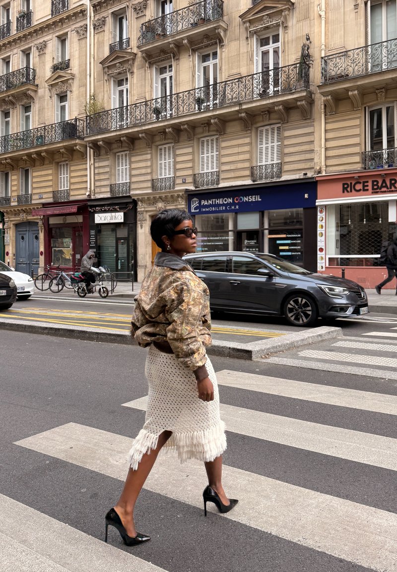 Femme portant des lunettes de soleil, une veste à motifs et une jupe blanche à franges marchant sur un passage pour piétons en ville, avec des bâtiments et des voitures en arrière-plan.