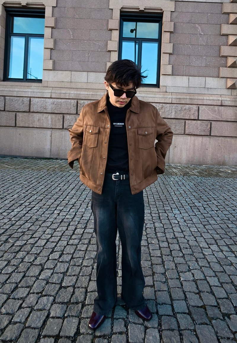 Jeune homme portant des lunettes de soleil, une veste marron, une chemise noire et un jean sombre, debout sur un pavé devant un bâtiment en pierre avec des fenêtres.