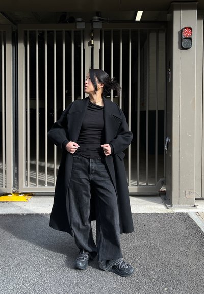 Abrigo de lana negro con solapas anchas sobre una camiseta negra ajustada, combinado con vaqueros negros holgados y zapatillas deportivas negras. Fondo de hormigón.