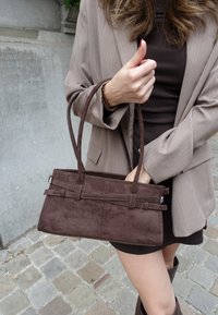 Kvinna i brun klänning och beige kavaj som håller en brun handväska och ger tumme upp på en kullerstensgatan.