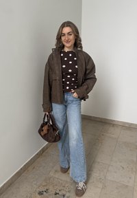 Jeune femme debout à l'intérieur portant une veste en cuir marron, un haut à pois, un jean bleu clair et tenant un sac à main marron avec un porte-clés en forme d'ours en peluche.