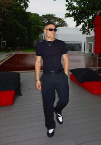 Camiseta preta ajustada, calças pretas soltas e sapatos preto e branco. Os acessórios incluem um colar de prata e óculos de sol escuros.