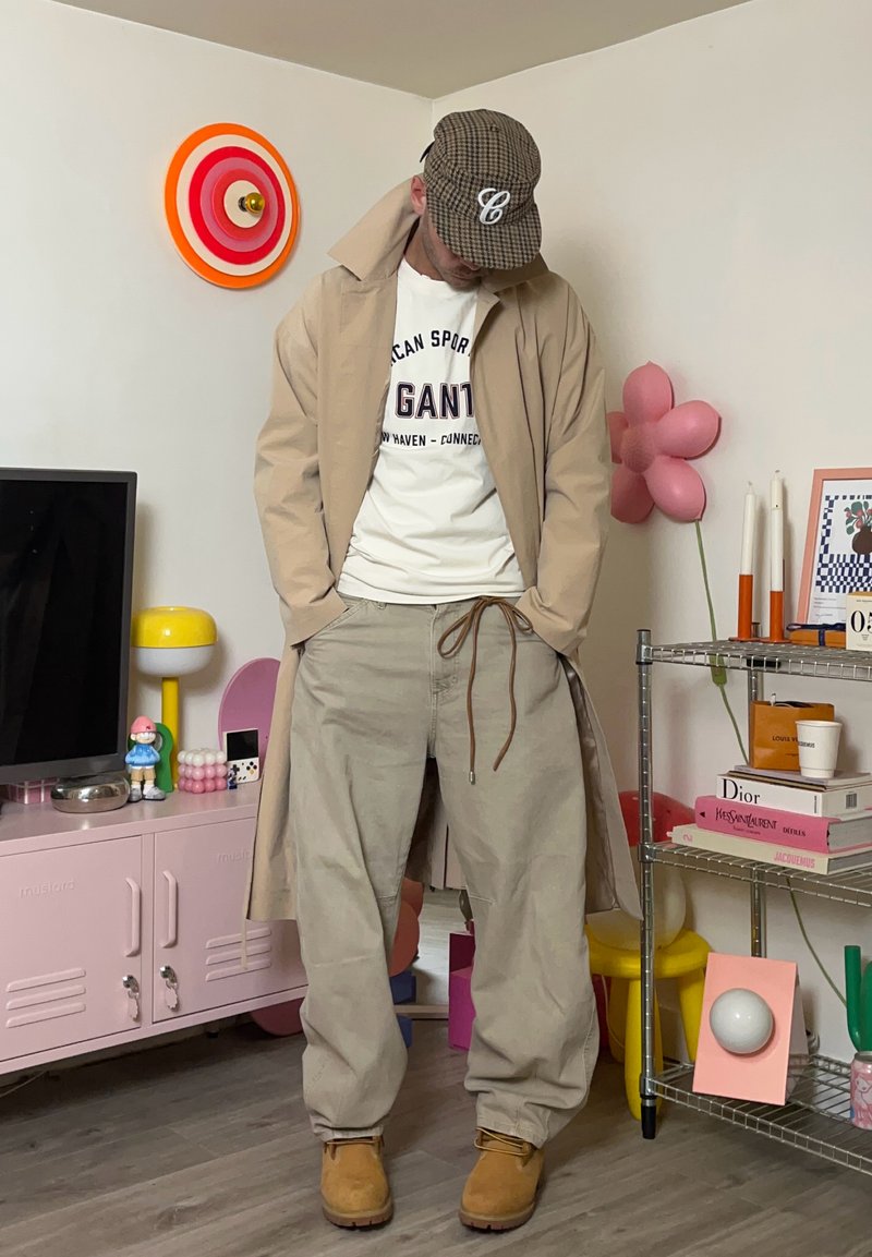 Trench beige, camicia bianca con stampa, pantaloni beige larghi, stivali marroni, berretto grigio a quadri. Decorazioni con fiori rosa e oggetti colorati sullo sfondo.