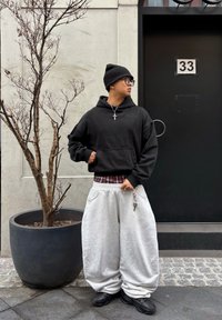 Jeune personne portant un bonnet noir, des lunettes, un sweat à capuche noir, un pantalon blanc ample avec un porte-clés, des chaussures noires, debout à côté d'un arbre en pot sans feuilles près d'une porte noire numérotée 33.