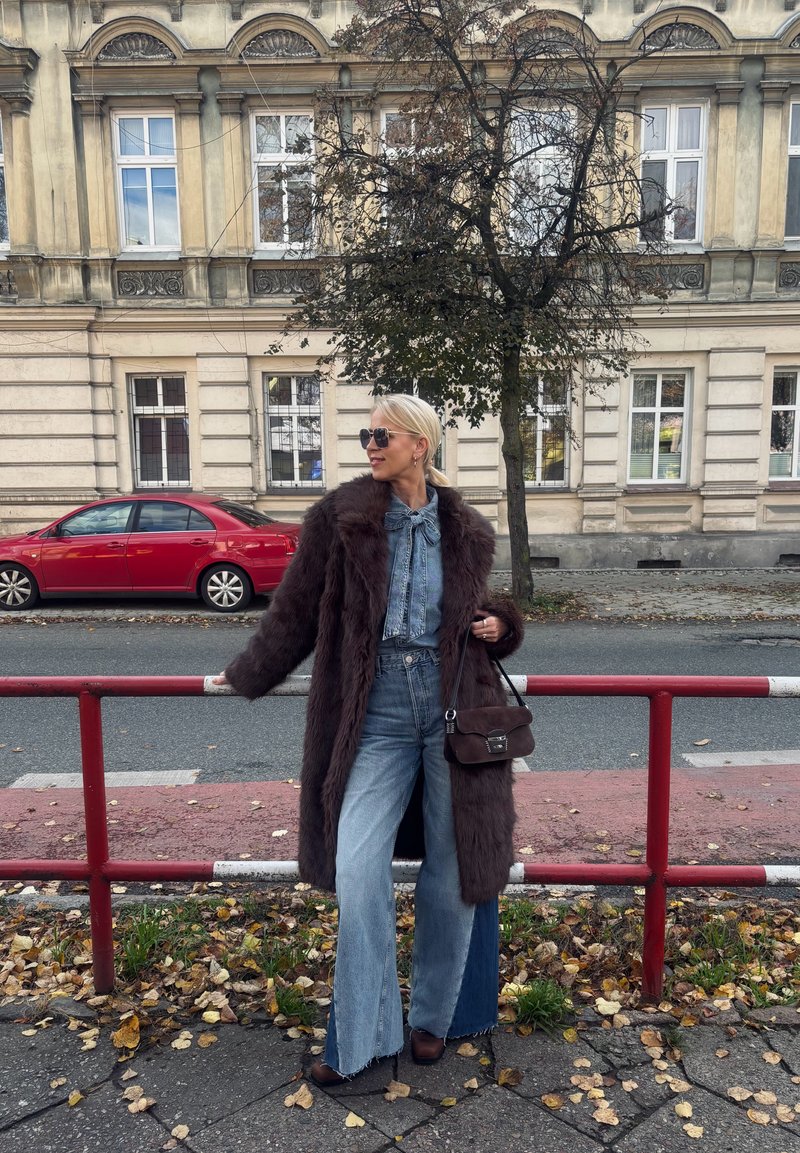 Brown faux fur coat over a denim shirt, wide-leg jeans, and dark brown heels. Accessories include sunglasses and a small brown handbag.