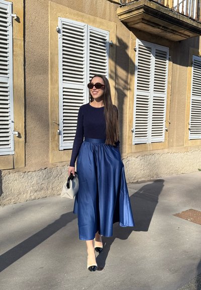 Falda midi de satén azul con cintura fruncida, combinada con una blusa ajustada de canalé azul marino. Los accesorios incluyen un bolso blanco pequeño y zapatos de tacón negros.