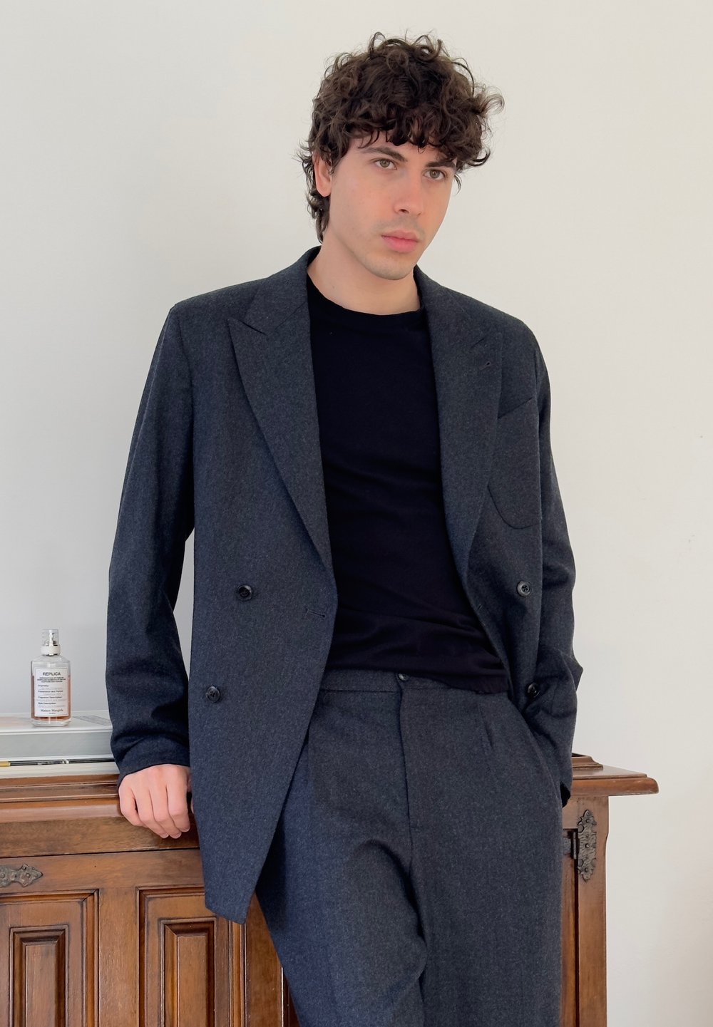 Junger Mann mit lockigem Haar, der einen dunkelgrauen Anzug und ein schwarzes Hemd trägt und sich an einem Holzschrank anlehnt, auf dem sich eine Parfümflasche befindet.