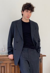 Junger Mann mit lockigem Haar, der einen dunkelgrauen Anzug und ein schwarzes Hemd trägt und sich an einem Holzschrank anlehnt, auf dem sich eine Parfümflasche befindet.