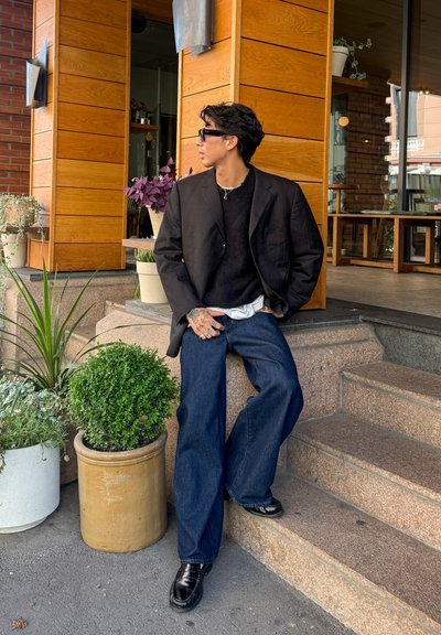 Blazer negro sobre un suéter negro, combinado con jeans anchos azules. Zapatos negros brillantes. Plantas en macetas y un exterior de madera en el fondo.