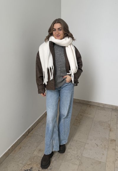 Mujer de pie en interiores con chaqueta marrón, suéter gris, bufanda blanca, jeans azul claro y botas negras, sonriendo a la cámara.