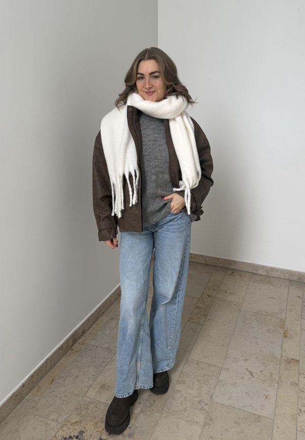 Woman standing indoors wearing a brown jacket, grey sweater, white scarf, light blue jeans, and black boots, smiling at the camera.