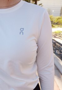 Weißes Langarmshirt mit strukturiertem Muster; verfügt über ein kleines graues Logo auf der linken Brustseite.