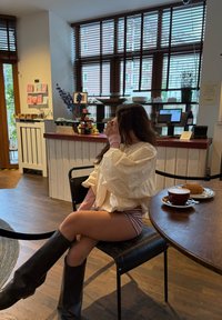 Chaqueta cream de gran tamaño, pantalones cortos a rayas rosas y botas negras hasta la rodilla, sentada en una mesa con una taza marrón y un pastel. Suelo de madera y grandes ventanales.