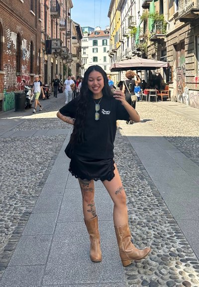 Una persona está de pie en una calle de adoquines vistiendo una camiseta negra y una falda negra con encaje, combinada con botas altas beige.