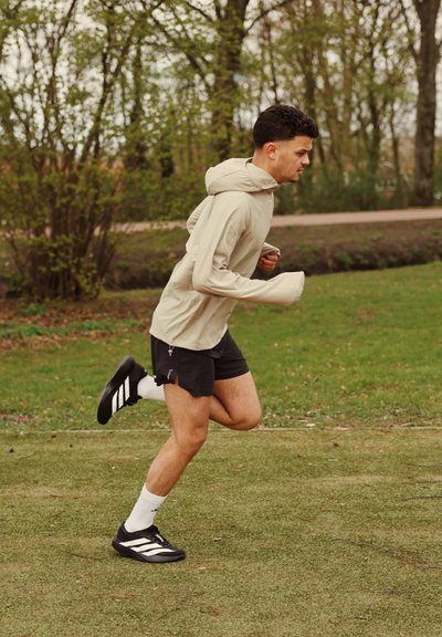 Hombre con sudadera beige y pantalones cortos negros corriendo en un sendero de césped en el parque, con árboles y arbustos al fondo.