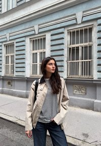 Béžová lehká bunda s černým límečkem, navlečená přes pruhované tričko, v kombinaci s tmavě modrými širokými džínami. Městské prostředí s texturovanými zdmi.