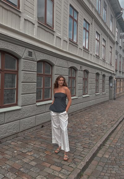 Top sin tirantes de color gris oscuro con un dobladillo tipo peplum, combinado con pantalones anchos blancos brillantes y sandalias marrones, contra una calle empedrada.