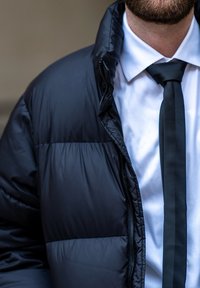 Fekete puffjaszerű dzseki sima, fényes textúrával, fehér ing és fekete nyakkendő fölött viselve, amely vatelin mintát és cipzár részleteket mutat.