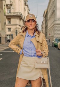 Manteau trench beige sur une chemise à rayures bleu clair et une mini-jupe crème. Les accessoires comprennent un sac à main de couleur claire et une casquette beige.