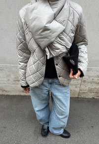 Personne portant une veste matelassée beige, un pull noir, un jean bleu clair et des chaussures noires, tenant un objet noir plié dans une main.