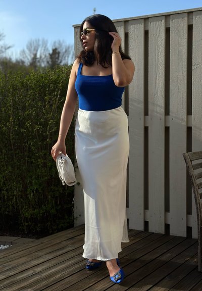 Top sin mangas azul ajustado, falda maxi de satén blanca, tacones azules con acentos decorativos, sosteniendo un bolso blanco, contra un fondo de una cubierta de madera y vegetación.