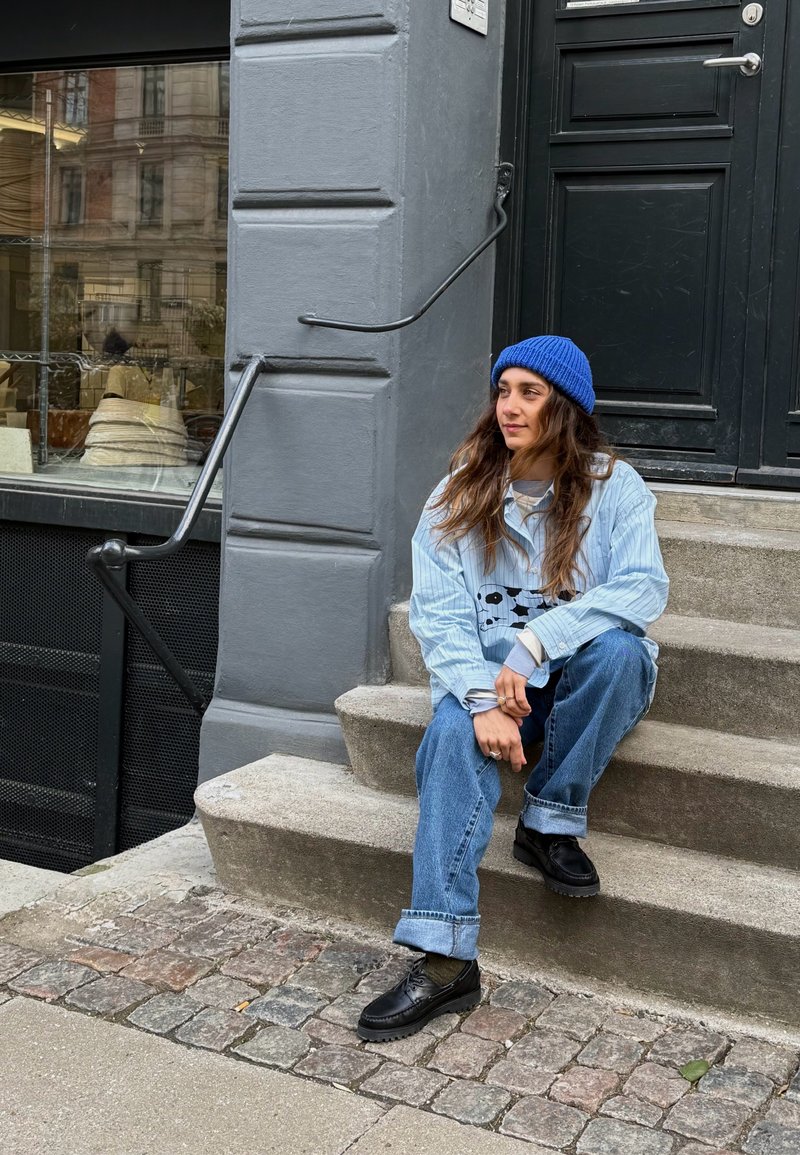 Νεαρή γυναίκα φορώντας μπλε σκουφί, ανοιχτό μπλε πουκάμισο και τζιν κάθεται στα σκαλιά από πέτρα σε εξωτερικό χώρο δίπλα σε σκούρα πόρτα, σε αστικό περιβάλλον.