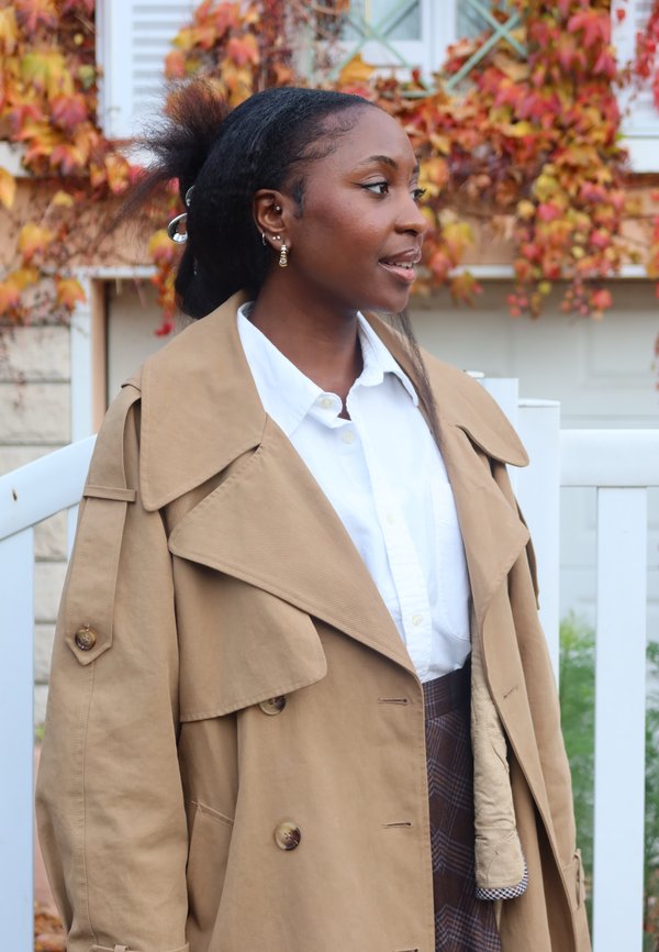 Beige trenchcoat met een brede kraag en grote knopen, gecombineerd met een wit overhemd. De textuur is glad, aangevuld met een rokje met patroon.