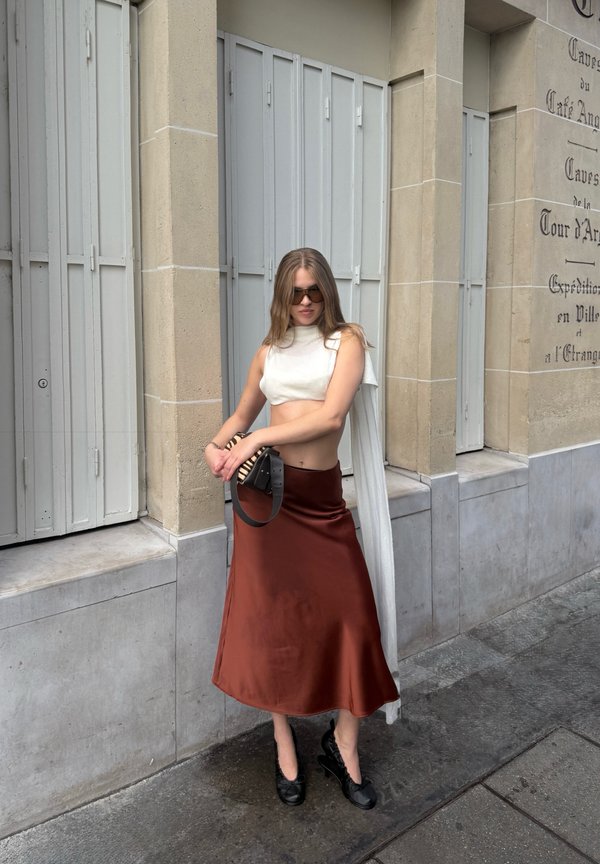 Joven mujer que lleva gafas de sol, top blanco corto, falda color óxido y zapatos negros, de pie en una acera de la ciudad cerca de un edificio beige.