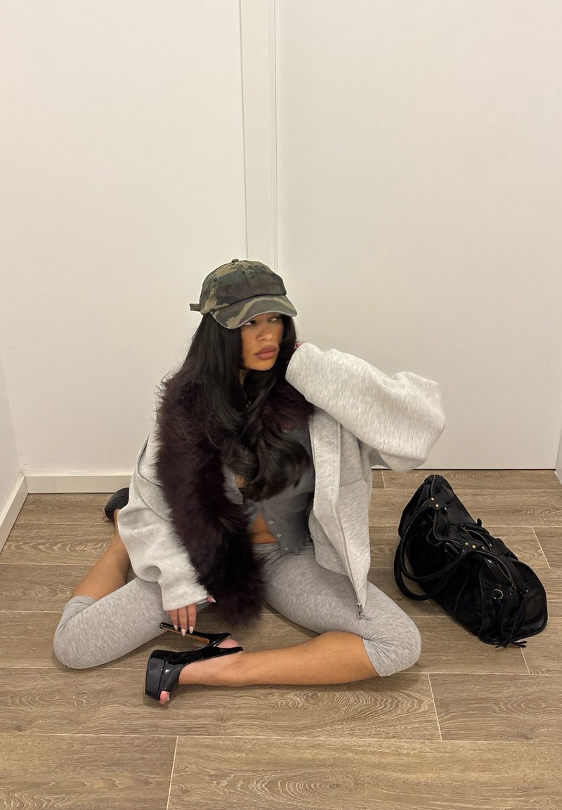 Jeune femme vêtue de vêtements décontractés gris, écharpe en fausse fourrure noire, casquette camouflage et chaussures à talons noires, assise sur un sol en bois à côté d'un sac à main noir contre un mur blanc.